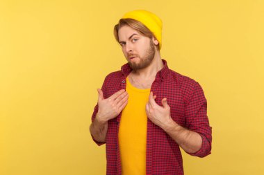 Şapkalı sakallı adam portresi ve damalı gömlek kameraya kibirli bakıyor, kendini işaret ediyor, zaferle övünüyor, kendinden emin. Sarı arka planda kapalı stüdyo çekimleri.