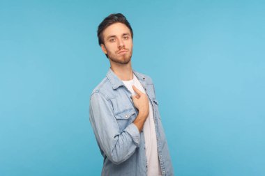 Portrait of man wearing denim shirt, pointing himself and looking with arrogant selfish expression, feeling successful and self-important. Indoor studio shot isolated on blue background. clipart
