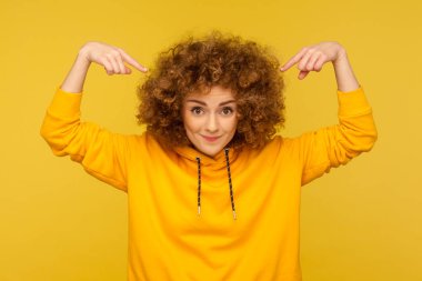 Kabarık kıvırcık saçlarına, kuaförüne ya da kuaförüne ya da kuaförüne, kuaförüne, makyaj ürünlerine, Afro saç stiline sahip bir kadına işaret eden bir kadın, günlük tarz kapüşonlu giyer. Sarı arka planda kapalı stüdyo çekimleri