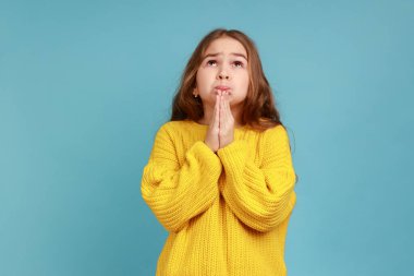 Küçük kızın portresi, yalvaran gözlerle Tanrı 'ya dua ederek çocukluk dilekleri diliyor ve umuyor sarı, sıradan bir süveter giyiyor. Mavi arka planda kapalı stüdyo çekimi.