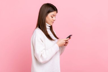 Akıllı telefon kullanan, sosyal ağları kontrol eden kadın portresi, beyaz günlük süveter giyen, konsantre bir ifadeye sahip. Pembe arka planda kapalı stüdyo çekimleri.