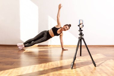 Sportif bir kadının yan kalas egzersizi videosu çekerken, tripoda telefon kamerası takarken, siyah spor bluz ve tayt giyerken yan görüntüsü. Pencereden gelen güneş ışığıyla aydınlatılan tam uzunluktaki stüdyo görüntüleri..