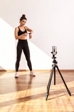 Ciddi bir atletik spor eğitmeni spor haberlerini canlı yayında sunuyor. Smartwatch 'i işaret ediyor. Siyah spor bluz ve tayt giyiyor. Pencereden gelen güneş ışığıyla aydınlatılan tam uzunluktaki stüdyo görüntüleri..