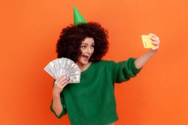 Afro saç stili, yeşil günlük süveter giyen ve elinde dolar tutan parti konisi ve canlı yayın yapan mutlu bir kadın. Turuncu arka planda kapalı stüdyo çekimleri.