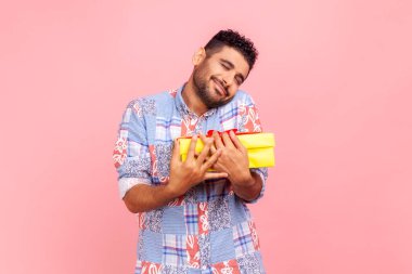 Genç, yakışıklı, sakallı, siyah saçlı, sarı paketli hediye kutusunu kucaklayan, hediye almaktan memnun, mavi gömlek giyen bir adam. Pembe arka planda kapalı stüdyo çekimleri.