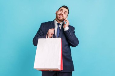 Elinde alışveriş poşetleri, elinde telefonla konuşan, büyük indirimlerle alışverişleriyle övünen, koyu renk ofis elbisesi giyen genç, mutlu bir adam. Mavi arka planda kapalı stüdyo çekimi.
