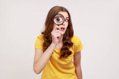 Portrait of funny young female in yellow casual T-shirt holding magnifying glass and looking at camera with big zoom eye, curious face. Indoor studio shot isolated on gray background. clipart