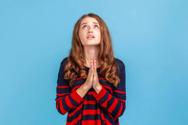 必死の猶予を求めて神に祈る 縞模様のカジュアルなスタイルのセーターを着て若い魅力的な女性の肖像画 青い背景に隔離された屋内スタジオショット — ストック写真