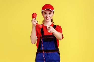 Elinde sabit telefonla kamerayı işaret eden, tulumlu ve kırmızı şapkalı gülümseyen pozitif işçi kadının portresi. Sarı arka planda kapalı stüdyo çekimleri.