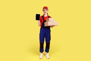 Kurye kadının tam boy portresi teslimat için paketler tutuyor ve reklam için boş ekranlı tableti gösteriyor, tulum ve şapka giyiyor. Sarı arka planda kapalı stüdyo çekimleri.