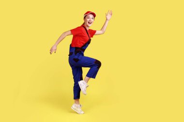 Bir bacağının üzerinde duran, kollarını kaldırmış, kameraya mutlu bir ifadeyle bakan, tulumlu ve kırmızı şapkalı heyecanlı bir kadının yan görünüm portresi. Sarı arka planda kapalı stüdyo çekimleri
