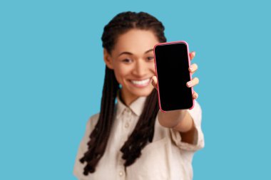 Siyah saçlı, iyimser bir kadın reklam ya da tanıtım için boş ekranlı, beyaz gömlekli akıllı bir telefon tutuyor. Mavi arka planda kapalı stüdyo çekimi.
