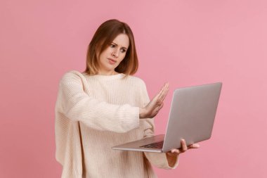 Dizüstü bilgisayara dur işareti gösteren bir kadın, video görüşmesinde konuşurken yasak işareti ile uyarı, online iletişim, beyaz kazak giyiyor. Pembe arka planda kapalı stüdyo çekimleri.