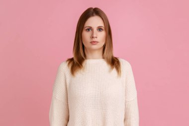 Çekici, otoriter, ciddi sarışın kadının portresi. Yüzünde sert bir ifadeyle kameraya bakıyor. Mutsuz, beyaz kazak giyiyor. Pembe arka planda kapalı stüdyo çekimleri.