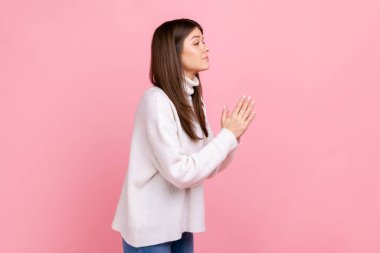 Kollarını dua jestinde tutan, af dileyen, üzgün hisseden, günlük beyaz süveter giyen güzel bir kızın yan görünümü. Pembe arka planda kapalı stüdyo çekimleri.