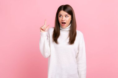 Heyecanlı esmer kadın parmak kaldırmış, mükemmel bir fikri ya da planı var, ağzını açık tutuyor, günlük beyaz süveter giyiyor. Pembe arka planda kapalı stüdyo çekimleri.