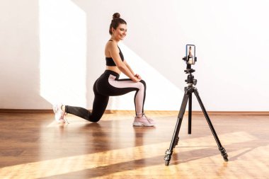 Çevrimiçi antrenör kadın bacak esnerken canlı yayın yapıyor, tripoda telefon kamerası takıyor, siyah spor bluz ve tayt giyiyor. Pencereden gelen güneş ışığıyla aydınlatılan tam uzunluktaki stüdyo görüntüleri..