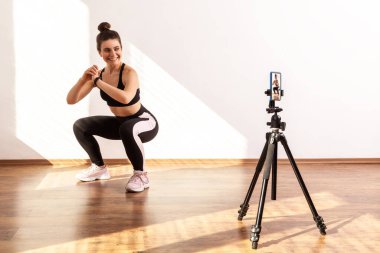 Gülümseyen koç doğru çömelme tekniğini öğretiyor, antrenman videosu kaydediyor, siyah spor bluzu ve tayt giyiyor. Pencereden gelen güneş ışığıyla aydınlatılan tam uzunluktaki stüdyo görüntüleri..