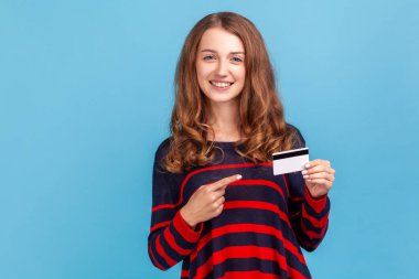 Çizgili bir süveter giyen, gülümseyen bir kadın, kredi kartına işaret eden, dişlek bir gülümsemeyle bakan, banka ya da alışveriş indirimi öneren. Mavi arka planda kapalı stüdyo çekimi.