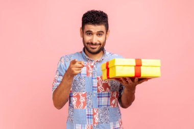Mavi tişört giyen, elinde hediye kutusu olan mutlu sakallı adam kamerayı gösteriyor, seni seçiyor, hediye veriyor, tatilini kutluyor. Pembe arka planda kapalı stüdyo çekimleri.