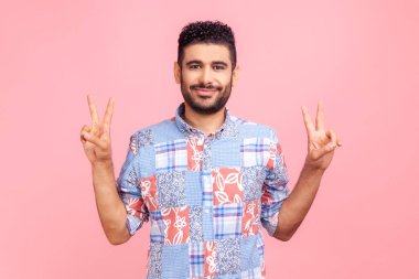 Mavi gömlekli, koyu renk saçlı ve sakallı mutlu adam zafer işaretini parmaklarıyla gösteriyor, kameraya gülümseyerek bakıyor. Pembe arka planda kapalı stüdyo çekimleri.