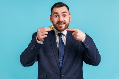 Kripto para birimine dikkat! Tatmin olmuş iş adamı resmi kıyafet giyiyor bitcoin doğrultuyor ve kameraya gülümsüyor, dijital para gösteriyor. Mavi arka planda kapalı stüdyo çekimi.