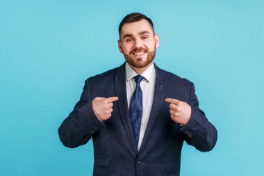 Ben geldim! Sakallı hırslı bir genç adam resmi tarz giyinmiş, yüzünde dişlek bir gülümsemeyle kendini işaret ediyor, kendine güvenen bir iş adamı. Mavi arka planda kapalı stüdyo çekimi.