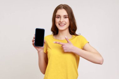 Cep telefonuma bak! Sevimli genç kız akıllı telefonu parmağıyla gösterip kameraya gülümsüyor ve mobil cihazın reklamını yapıyor. Gri arka planda izole edilmiş kapalı stüdyo görüntüleri.