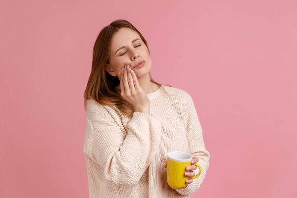 Portrait of unhealthy ill blond woman felling toothache after drinking hot or cold beverage, holding cup in hand, wearing white sweater. Indoor studio shot isolated on pink background.
