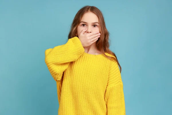少女の手で口をカバーの肖像画 秘密を言うことを恐れて 子供は話すことを恐れて 黄色のカジュアルなスタイルのセーターを着ている 青い背景に隔離された屋内スタジオショット — ストック写真