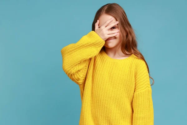 女の子の手で顔をカバーし 指を見て 好奇心と恥ずかしがり屋の秘密を見て 黄色のカジュアルなスタイルのセーターを着ている 青い背景に隔離された屋内スタジオショット — ストック写真