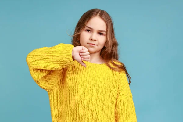 かわいい女の子の肖像画眉をひそめ 親指をジェスチャー 否定的な不承認のサインを示し 黄色のカジュアルなスタイルのセーターを着て 青い背景に隔離された屋内スタジオショット — ストック写真