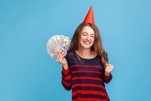縞のカジュアルなスタイルのセーターやパーティーコーンを着て幸せな女性は ドル札を保持し 誕生日を祝い 贈り物としてお金を得ることを喜んでいます 青い背景に隔離された屋内スタジオショット — ストック写真
