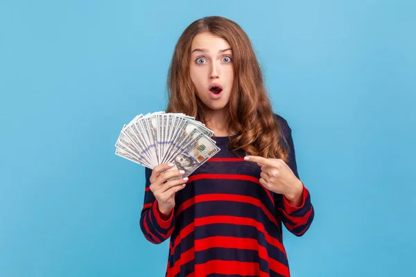 縞模様のカジュアルなスタイルのセーターを着てショックを受けた女性は 彼女の手の中にドル紙幣を指して 大きな利益を驚かせた 青い背景に隔離された屋内スタジオショット — ストック写真