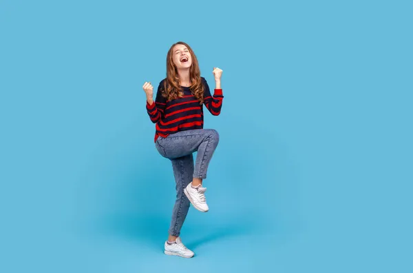 縞模様のカジュアルなスタイルのセーターとジーンズを着て 完全な長さ正の満足女性は 勝利を表現し 勝利の幸せされて 握りしめ拳を調達した 青い背景に隔離された屋内スタジオショット — ストック写真