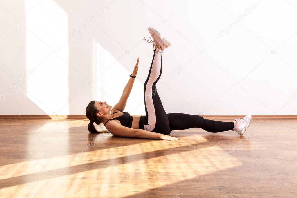 Sportive female raising hand and leg, doing crunches, pumping up abdominal and spine muscles, wearing black sports top and tights. Full length studio shot illuminated by sunlight from window.