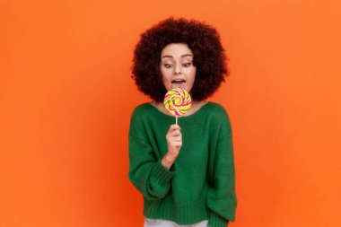 Afro saç stili olan, yeşil, gündelik süveter giyen, çok renkli şeker yiyen, yemek yemek isteyen, lolipopa bakmak isteyen çekici bir kadın. Turuncu arka planda kapalı stüdyo çekimleri.