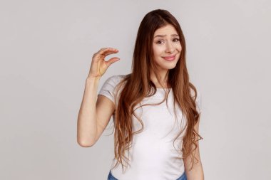 Koyu renk saçlı mutsuz bir kadının portresi. Hayal kırıklığına uğramış yüz ifadesi olan küçük bir jest. Beyaz tişört giyiyor. Gri arka planda izole edilmiş kapalı stüdyo görüntüleri.