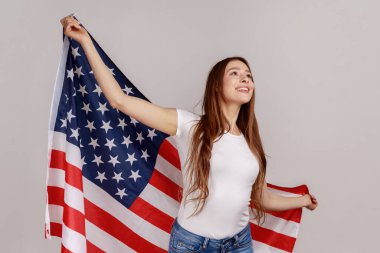 Siyah saçlı mutlu bir kadının portresi, elinde Amerikan bayrağı, başka tarafa bakıyor, ulusal bayramı kutluyor, beyaz tişört giyiyor. Gri arka planda izole edilmiş kapalı stüdyo görüntüleri.