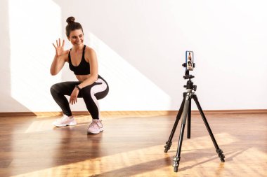 Koç, fitness vlog 'u için video kaydediyor, takipçilerle takılıyor, kalan sayıyı gösteriyor, siyah spor bluzu ve tayt giyiyor. Pencereden gelen güneş ışığıyla aydınlatılan tam uzunluktaki stüdyo görüntüleri..