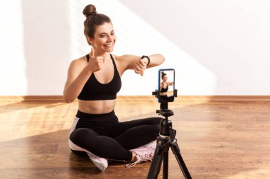 Spor eğitmeni çevrimiçi antrenman yapıyor, sonuçları takip cihazıyla kontrol ediyor, baş parmağını gösteriyor, siyah spor bluzu ve tayt giyiyor. Pencereden gelen güneş ışığıyla aydınlatılan tam uzunluktaki stüdyo görüntüleri..