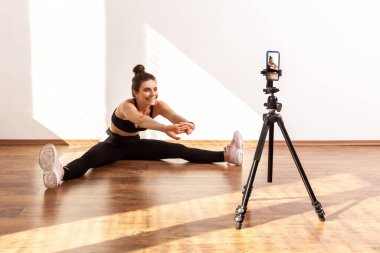 Spor salonundan sonra esneme dersleri almak için tripodun üzerinde telefon kullanan siyah spor bluzu ve tayt giyen bir bayan antrenör. Pencereden gelen güneş ışığıyla aydınlatılan tam uzunluktaki stüdyo görüntüleri..
