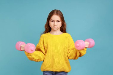 Elinde pembe dambıllar tutan küçük bir kızın portresi, ciddi bir ifadeyle kameraya bakıyor, sarı günlük süveter giyiyor. Mavi arka planda kapalı stüdyo çekimi.