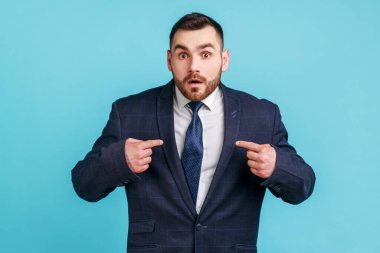 Vay canına, bu benim! Koyu takım elbiseli, hayretler içinde kameraya bakıp kendini gösteren ve kişisel başarısından şok olan şaşkın iş adamının portresi. Mavi arka planda kapalı stüdyo çekimi.
