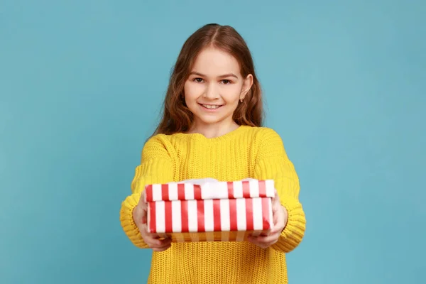 女の子の笑顔でカメラにギフトボックスを与える 休日に子供の挨拶やプレゼントを共有し 黄色のカジュアルなスタイルのセーターを着て 青い背景に隔離された屋内スタジオショット — ストック写真