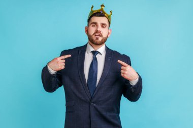 Resmi tarz kıyafet giymiş gururlu sakallı bir adam kameraya egoist bir şekilde bakıyor, başında tacıyla poz veriyor, kral gibi davranıyor, kendini işaret ediyor. Mavi arka planda kapalı stüdyo çekimi.