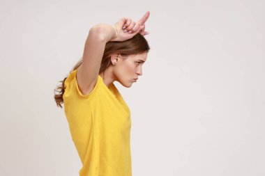 İş yerinde anlaşmazlık. Saldırgan genç bir kadın profili, kahverengi saçlı, sıradan giyinmiş, boğa boynuzu hareketi yapan, saldırmakla tehdit eden. Gri arka planda izole edilmiş kapalı stüdyo görüntüleri.