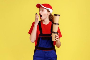 Uykulu yorgun işçi kadın elinde tek kullanımlık kahve fincanları, esniyor, ağzı elle kapanıyor, iş üniforması ve şapka giyiyor. Sarı arka planda kapalı stüdyo çekimleri.
