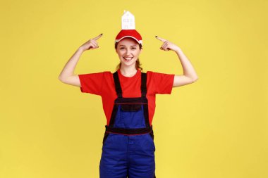 Kameraya bakan mutlu iyimser kadın portresi, kafasındaki kağıt evi gösteren, iş üniforması giyen ve kırmızı şapkalı. Sarı arka planda kapalı stüdyo çekimleri.