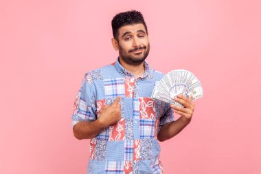 Sakallı ve hoş görünümlü genç bir adam elinde dolarlarla gururla mavi bir gömlek giyerek kendini işaret ediyor. Pembe arka planda kapalı stüdyo çekimleri.